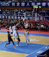 A basketball game takes place in a large indoor arena filled with spectators. Players from two teams, one in black uniforms and the other in white, compete for the ball near the hoop. The stadium is adorned with banners in Chinese, and a scoreboard displays a tied score of 21. The court features a distinctive blue and wood pattern.