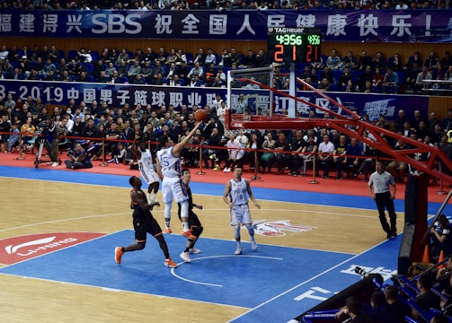 A basketball game takes place in a large indoor arena filled with spectators. Players from two teams, one in black uniforms and the other in white, compete for the ball near the hoop. The stadium is adorned with banners in Chinese, and a scoreboard displays a tied score of 21. The court features a distinctive blue and wood pattern.