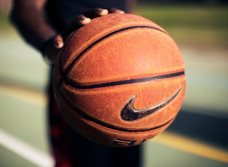 A detailed close-up of a hand holding a well-worn basketball with the Nike logo in focus. The blurred background suggests an outdoor basketball court with green and yellow hues, indicating lines and potential grassy areas.