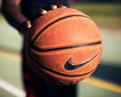 A detailed close-up of a hand holding a well-worn basketball with the Nike logo in focus. The blurred background suggests an outdoor basketball court with green and yellow hues, indicating lines and potential grassy areas.