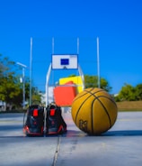 A basketball and a pair of black sports shoes are placed on an outdoor basketball court. A basketball hoop with a backboard is visible in the background, and the court is surrounded by trees and some buildings under a clear blue sky.