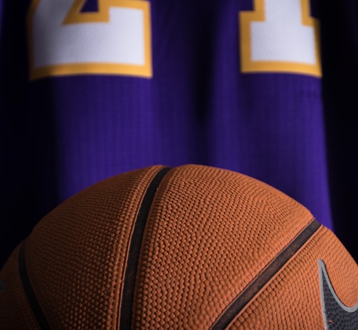A close-up view of a textured basketball positioned in front of a purple sports jersey with the number 24 in white and yellow.