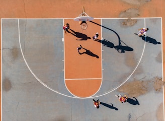A top-down view of a basketball court with several players engaged in a game. The court features a distinctive orange and blue color scheme, and the shadows cast by the players suggest a sunny day. The basketball hoop, lines of the court, and multiple players in motion are clearly visible.