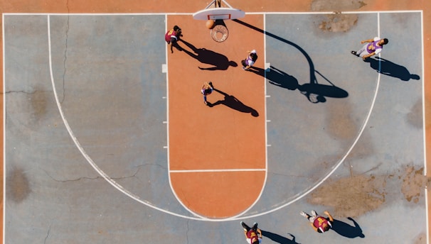 A top-down view of a basketball court with several players engaged in a game. The court features a distinctive orange and blue color scheme, and the shadows cast by the players suggest a sunny day. The basketball hoop, lines of the court, and multiple players in motion are clearly visible.