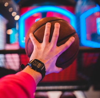 A hand wears a digital watch and holds a basketball, aiming towards a basketball hoop with vibrant neon lights in the background, creating a dynamic and energetic atmosphere.