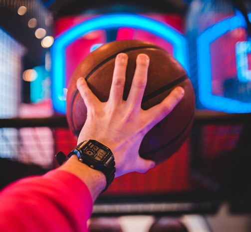 A hand wears a digital watch and holds a basketball, aiming towards a basketball hoop with vibrant neon lights in the background, creating a dynamic and energetic atmosphere.