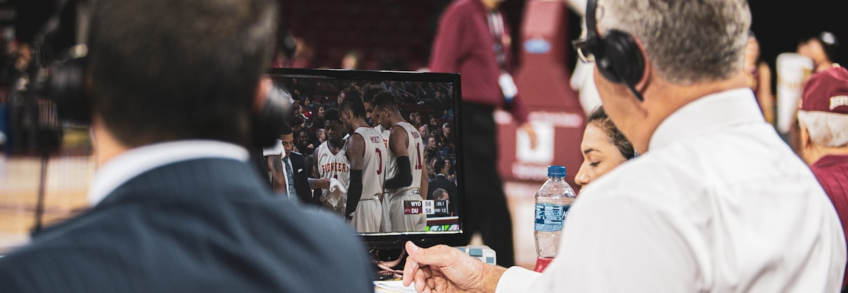 Two men wearing headsets focus on a monitor displaying a basketball team in discussion. Others can be seen around them, including a woman with a bottle of water, and an ongoing basketball game in the background.