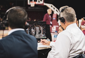 Two men wearing headsets focus on a monitor displaying a basketball team in discussion. Others can be seen around them, including a woman with a bottle of water, and an ongoing basketball game in the background.