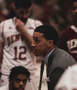 A basketball coach is in an intense discussion with his team. He is wearing a gray suit and tie, leaning forward with a focused expression. The players surrounding him are in white and maroon jerseys with 'Denver' printed on them. One player is sitting and listening attentively while another stands with a pensive look.