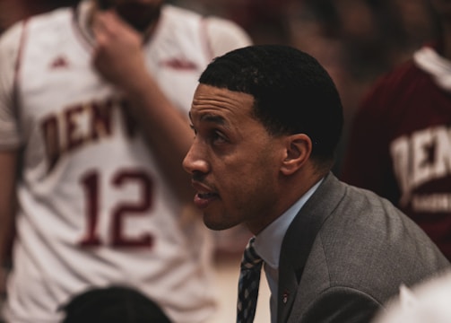A basketball coach is in an intense discussion with his team. He is wearing a gray suit and tie, leaning forward with a focused expression. The players surrounding him are in white and maroon jerseys with 'Denver' printed on them. One player is sitting and listening attentively while another stands with a pensive look.