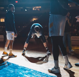 Basketball players are on a court, engaging in what appears to be a warm-up or team introduction session. The lighting is dramatic, casting strong shadows on the wooden floor. A player in the middle is stretching while others stand around him in sports attire.