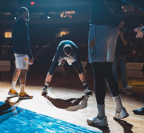 Basketball players are on a court, engaging in what appears to be a warm-up or team introduction session. The lighting is dramatic, casting strong shadows on the wooden floor. A player in the middle is stretching while others stand around him in sports attire.