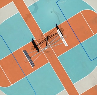 An aerial view of a basketball court with distinctive orange and blue sections. Multiple basketball hoops are positioned at different angles, casting shadows on the ground.