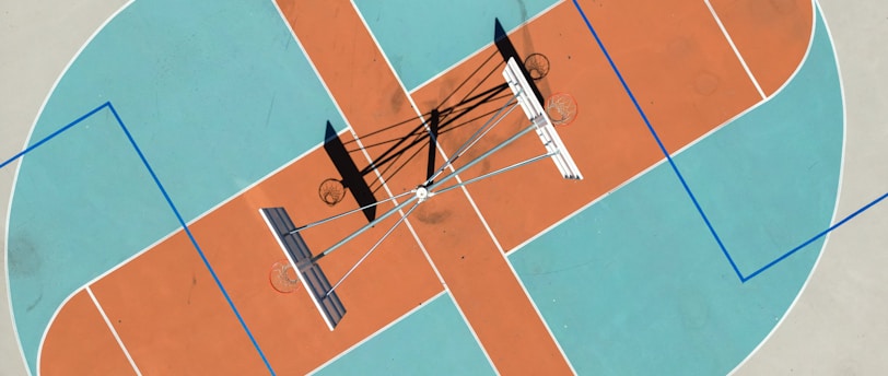 An aerial view of a basketball court with distinctive orange and blue sections. Multiple basketball hoops are positioned at different angles, casting shadows on the ground.