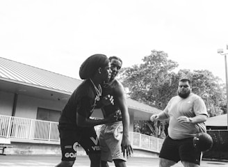 Three people are engaged in a basketball game on an outdoor court. Two individuals are closely interacting, possibly defending or strategizing, while the third person stands nearby holding a basketball. The perspective includes a close-up view of the basketball hoop above and some surrounding trees and buildings in the background.