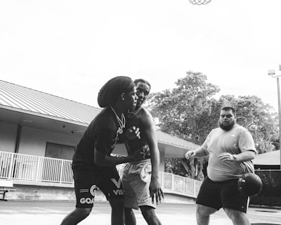 Three people are engaged in a basketball game on an outdoor court. Two individuals are closely interacting, possibly defending or strategizing, while the third person stands nearby holding a basketball. The perspective includes a close-up view of the basketball hoop above and some surrounding trees and buildings in the background.