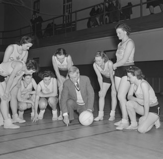A group of female basketball players in uniforms gathers around a coach who is crouching near a basketball. They appear to be strategizing or planning, with the coach pointing at the court. The background includes a balcony with people observing the scene.