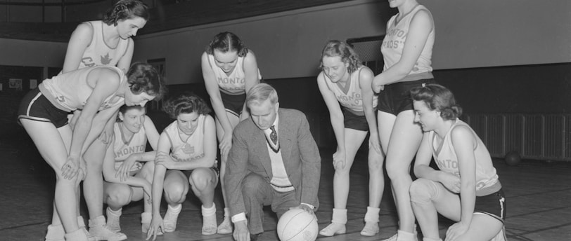 A group of female basketball players in uniforms gathers around a coach who is crouching near a basketball. They appear to be strategizing or planning, with the coach pointing at the court. The background includes a balcony with people observing the scene.