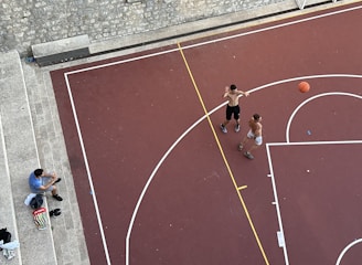 An aerial view of a basketball court with two shirtless individuals playing basketball. A basketball is in motion, with one player attempting a shot. Another person, sitting on stone steps nearby, appears to be spectating or taking a break. Various personal items, including a bag and shoes, are placed on the steps.