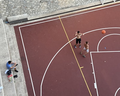 An aerial view of a basketball court with two shirtless individuals playing basketball. A basketball is in motion, with one player attempting a shot. Another person, sitting on stone steps nearby, appears to be spectating or taking a break. Various personal items, including a bag and shoes, are placed on the steps.