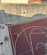 An aerial view of an outdoor basketball court with two people playing basketball. The court has clearly marked lines and a hoop with a backboard. Three individuals are sitting on the bleachers to the side of the court, facing towards the game. The court is bordered by a stone wall with a small window, casting a shadow on part of the court.