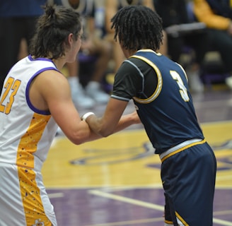 Two basketball players are interacting on a court, with a focus on their jerseys numbered 23 and 3. The players are in motion, engaged in a dynamic play. In the background, other team members sit on the benches dressed in matching uniforms, with blurred figures suggesting spectators or coaching staff.