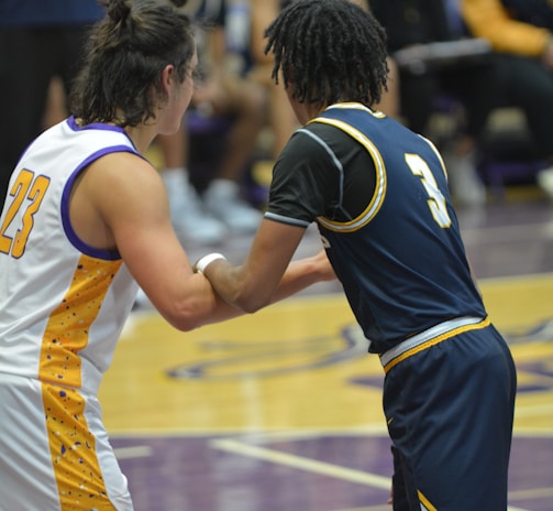 Two basketball players are interacting on a court, with a focus on their jerseys numbered 23 and 3. The players are in motion, engaged in a dynamic play. In the background, other team members sit on the benches dressed in matching uniforms, with blurred figures suggesting spectators or coaching staff.